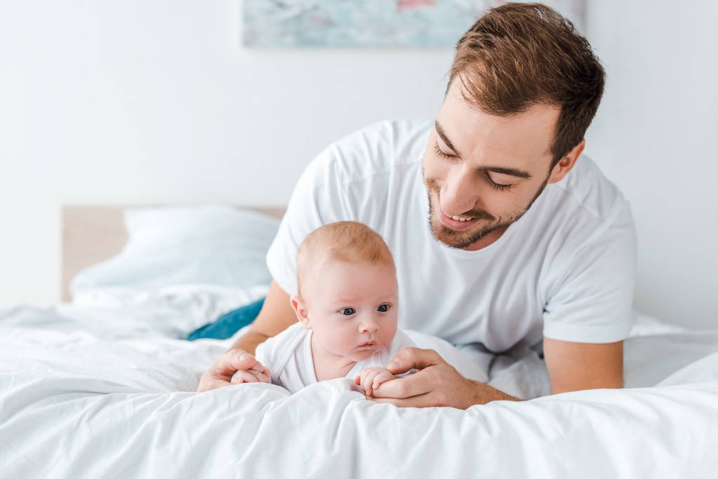 Sonriente padre acostado en la cama con el bebé en el dormitorio
 - Foto, Imagen