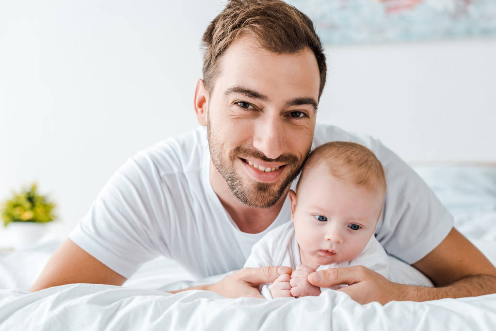 Sonriente padre acostado en la cama con el bebé en el dormitorio
 - Foto, Imagen