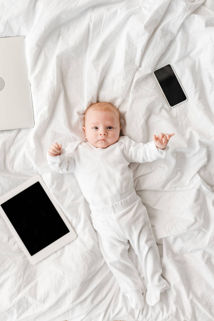 bovenaanzicht van de baby liggend op bed met laptop, digitale Tablet en smartphone met blanco scherm - Foto, afbeelding