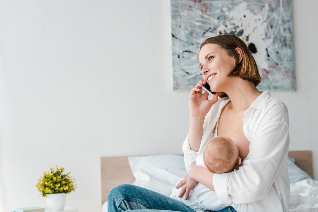 Lachende vrouw praten op smartphone terwijl borstvoeding baby thuis - Foto, afbeelding