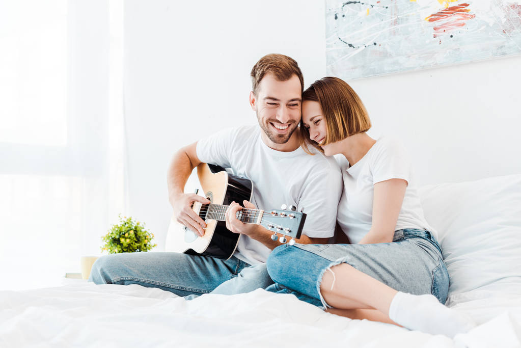 Χαμογελαστός άνθρωπος καθισμένος στο κρεβάτι με τη γυναίκα του και παίζοντας κιθάρα - Φωτογραφία, εικόνα