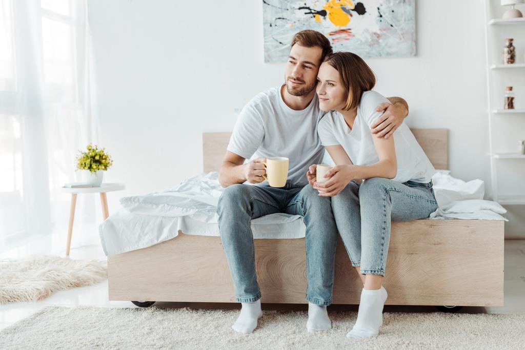 couple with cups of coffee sitting on bed and looking away - Photo, Image