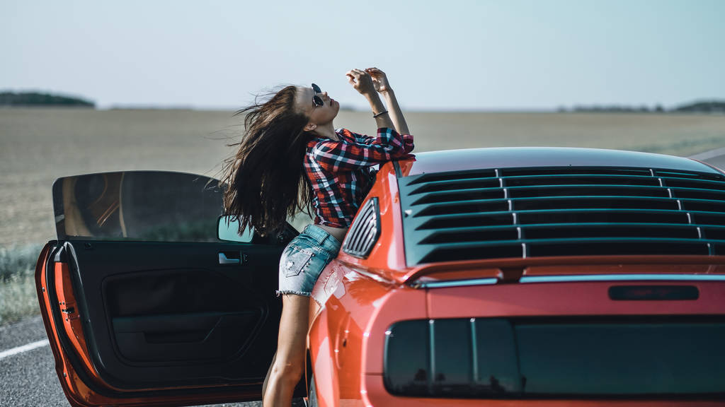 linda menina morena vestida com shorts curtos e uma camisa com óculos de sol perto de um carro esportivo vermelho
 - Foto, Imagem