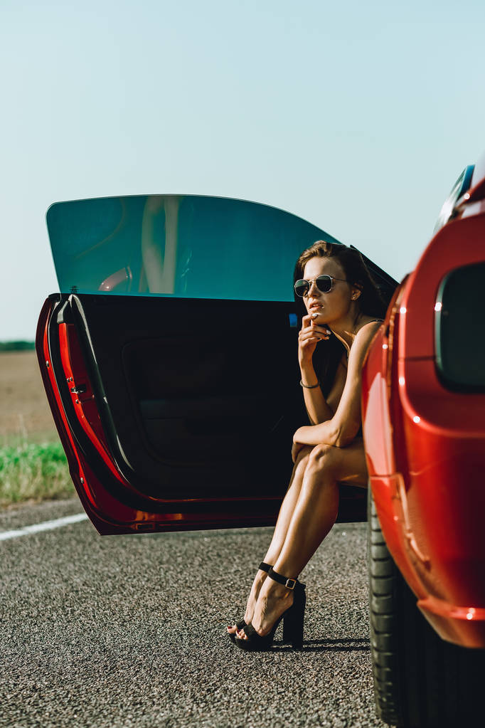 Piękna dziewczyna w Body z długimi włosami i okularami przeciwsłonecznymi siedzi na kapturze czerwonego samochodu, na drodze - Zdjęcie, obraz