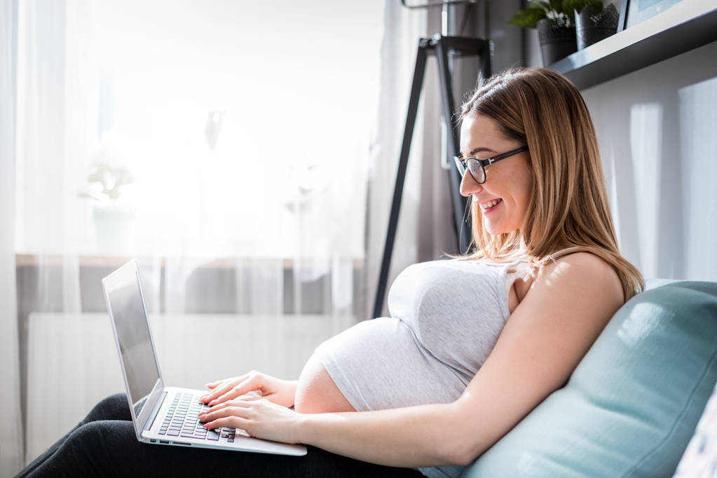 Χαμογελαστή έγκυος γυναίκα χρησιμοποιώντας φορητό υπολογιστή, κάθεται στον καναπέ στο σπίτι - Φωτογραφία, εικόνα