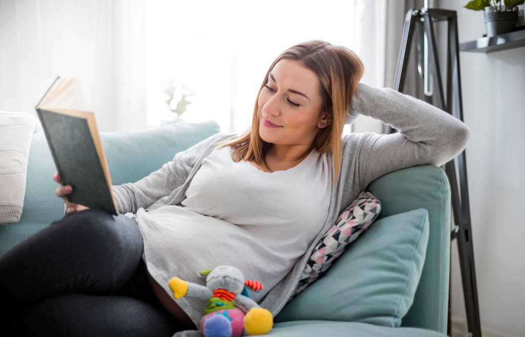 Έγκυος γυναίκα χαλαρωτικό στον καναπέ διαβάζοντας το βιβλίο κοιτάζοντας την κοιλιά - Φωτογραφία, εικόνα