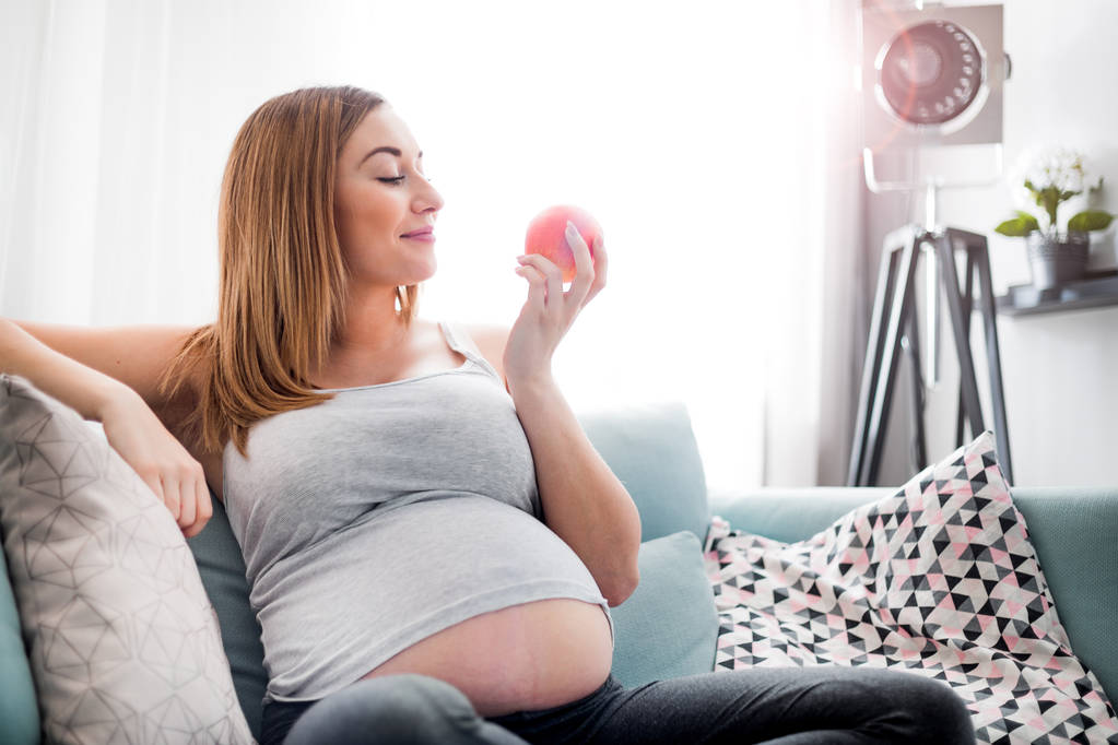 Těhotná žena jedla jablko a odpočívala na pohovce doma - Fotografie, Obrázek