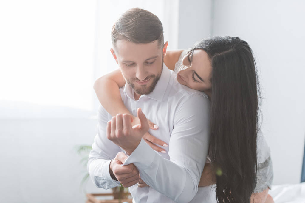 cheerful girlfriend hugging bearded boyfriend in white shirt at home  - Photo, Image