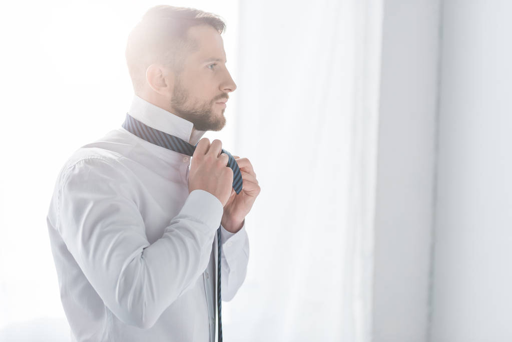 επιτυχημένος άντρας με λευκό πουκάμισο αγγίζοντας γραβάτα, ενώ στέκεται στο σπίτι  - Φωτογραφία, εικόνα