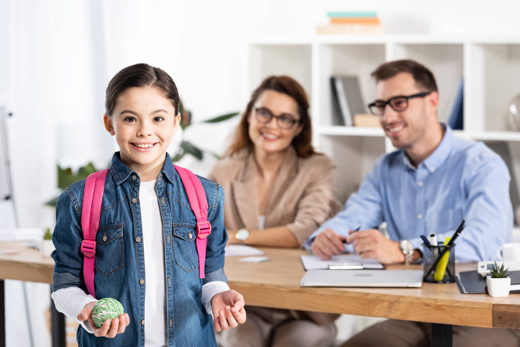 selective focus of cheerful kid with backpack holding ball near happy parents in office  - Photo, Image