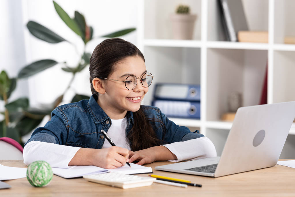 niño feliz mirando el ordenador portátil mientras sostiene la pluma cerca del cuaderno en la oficina
  - Foto, imagen