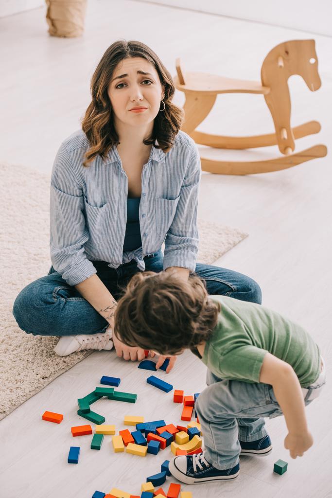 λυπημένη κουρασμένη μητέρα κάθεται στο πάτωμα, ενώ ο γιος παίζοντας με μπλοκ παιχνιδιών - Φωτογραφία, εικόνα