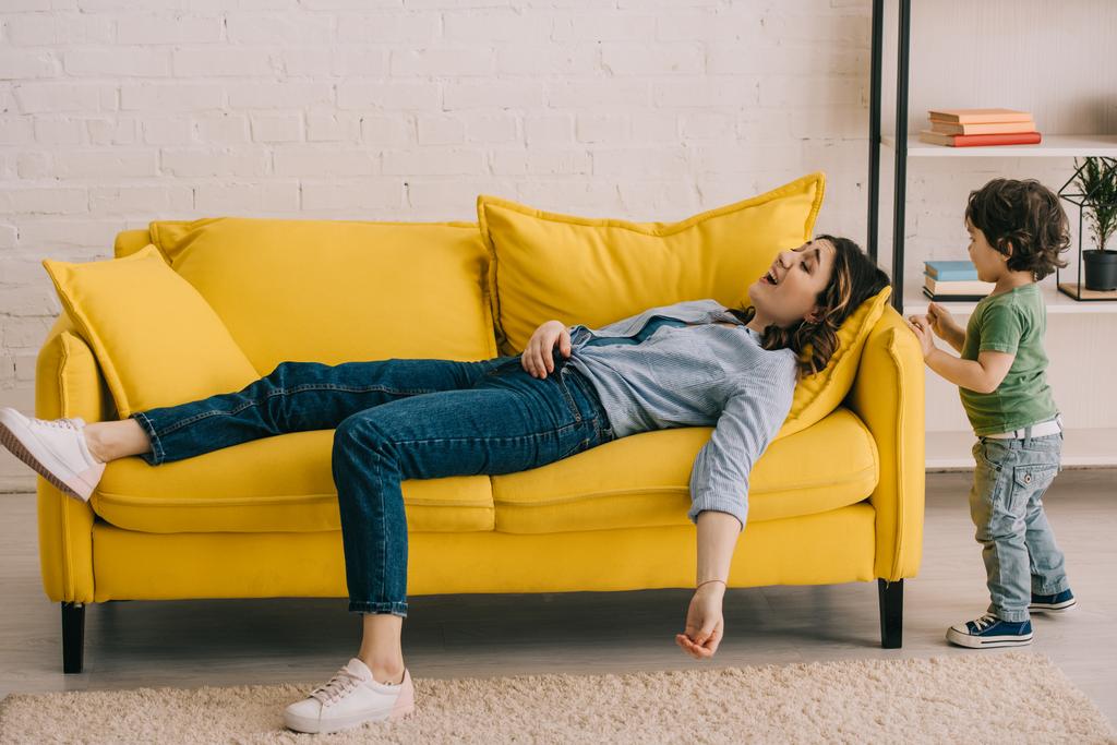 menino de pé perto da mãe cansada deitado no sofá amarelo na sala de estar
 - Foto, Imagem