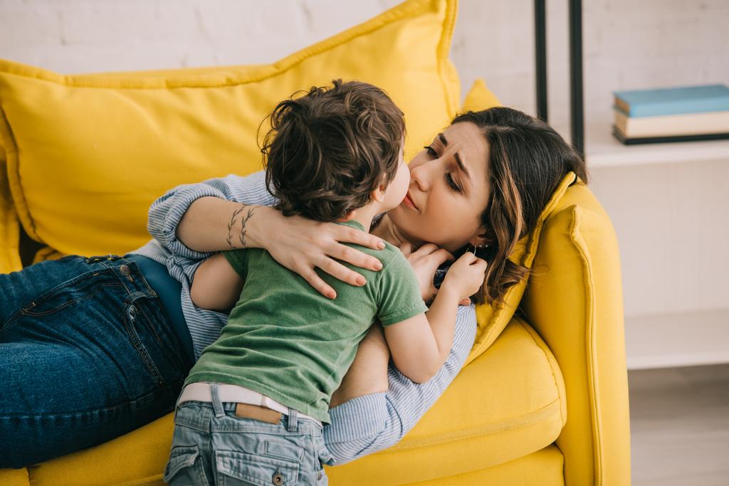 Little boy kissing tired mother lying on yellow sofa - Photo, Image