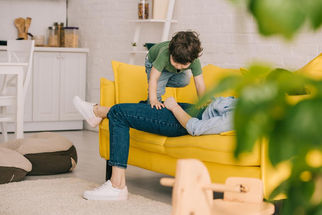 μικρό αγόρι που παίζει με την κουρασμένη μητέρα, ενώ αυτή κείτεται σε κίτρινο καναπέ στο σαλόνι - Φωτογραφία, εικόνα
