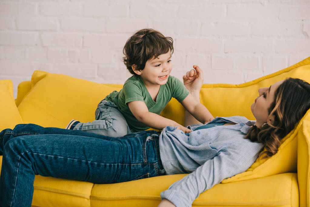 bambino seduto su madre stanca mentre lei sdraiata sul divano giallo in soggiorno
 - Foto, immagini