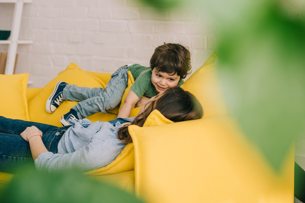 little boy playing with tired mother while she lying on yellow sofa in living room - Photo, Image