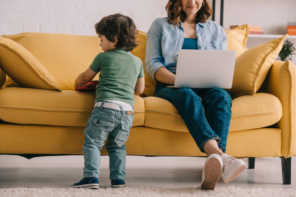 ребенок стоит рядом с диваном в то время как мать работает с ноутбуком
 - Фото, изображение