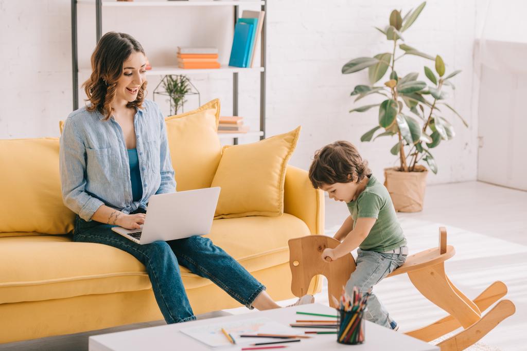kid sitting on rocking horse while mother working with laptop in living room - Photo, Image