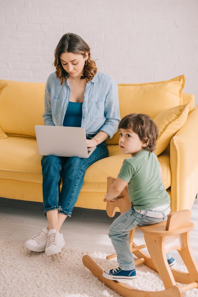 kind zittend op Rocking Horse terwijl moeder werken met laptop in de woonkamer - Foto, afbeelding