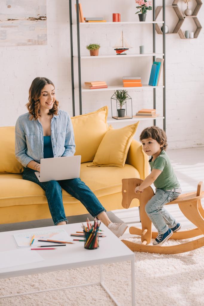 Kind sitzt auf Schaukelpferd, während Mutter im Wohnzimmer mit Laptop arbeitet - Foto, Bild