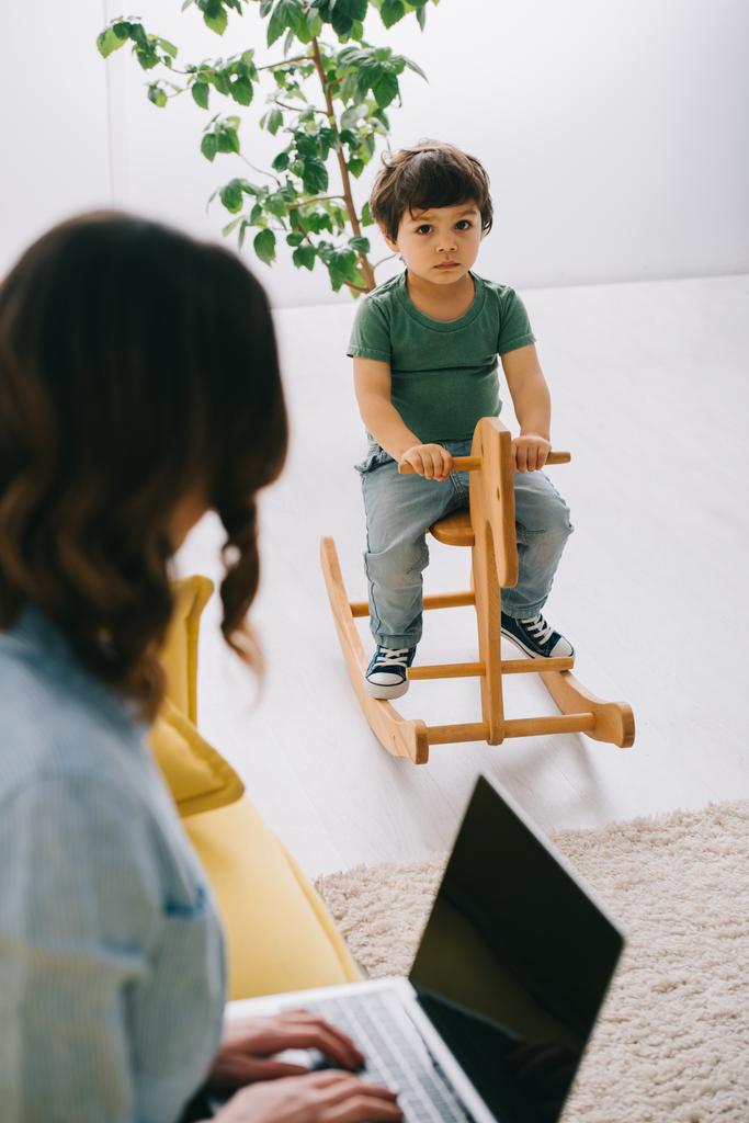 kid sitting on rocking horse while mother working with laptop in living room - Photo, Image