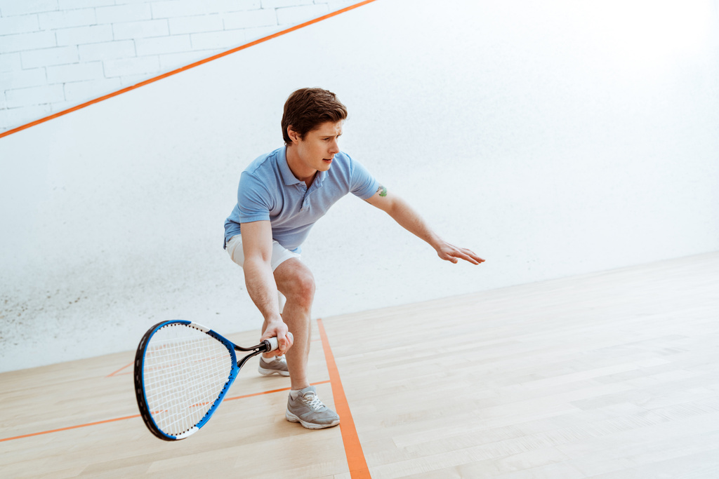Tunteellinen urheilija sinisessä poolopaidassa pelaamassa squashia neliseinäisessä hovissa
 - Valokuva, kuva