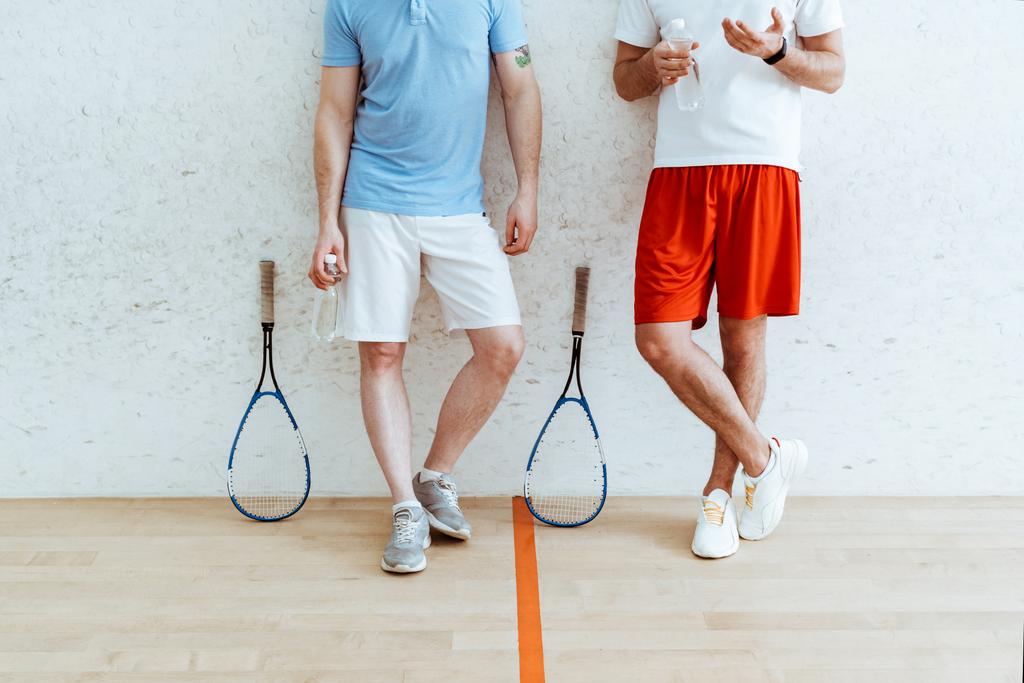 Vista ritagliata di due giocatori di squash in pantaloncini in piedi con gambe incrociate in un campo a quattro pareti
 - Foto, immagini