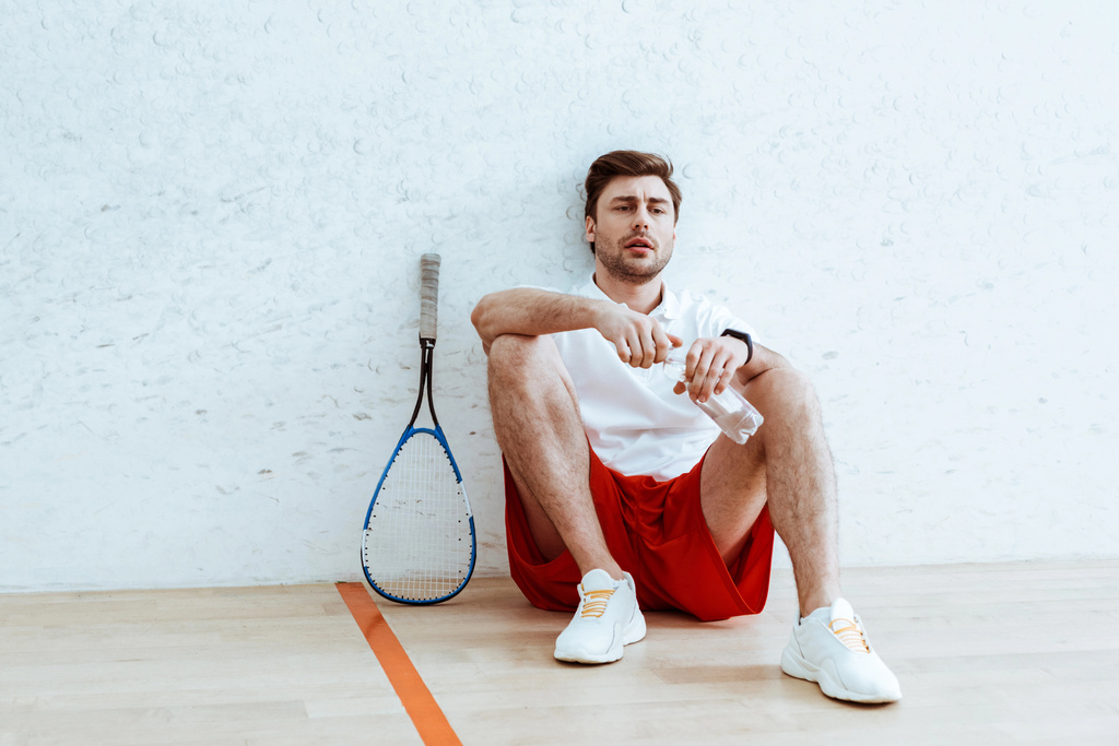 Cansado jogador squash sentado no chão e abertura garrafa de água
 - Foto, Imagem