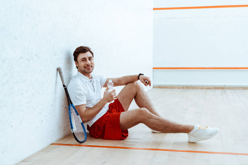 Squash-Spieler sitzt am Boden und hält eine Flasche Wasser in der Hand - Foto, Bild