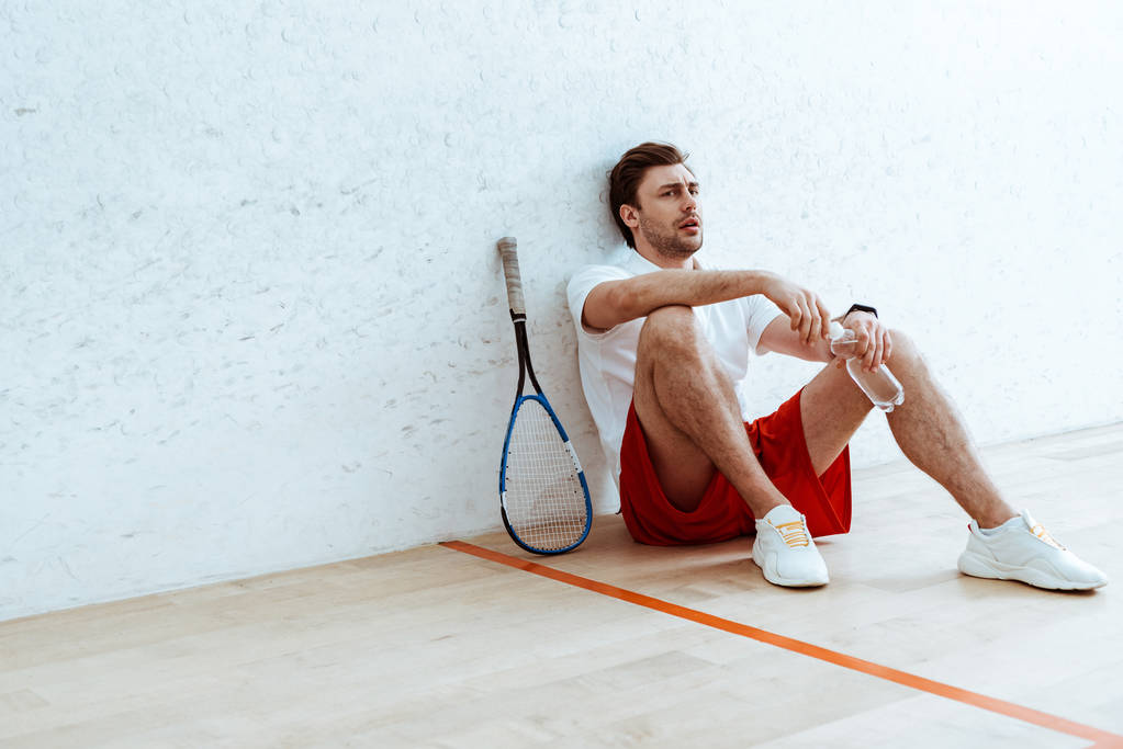 Squash-Spieler sitzt am Boden und hält eine Flasche Wasser in der Hand - Foto, Bild