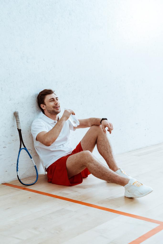 Jogador de squash em shorts vermelhos sentado no chão e água potável
 - Foto, Imagem