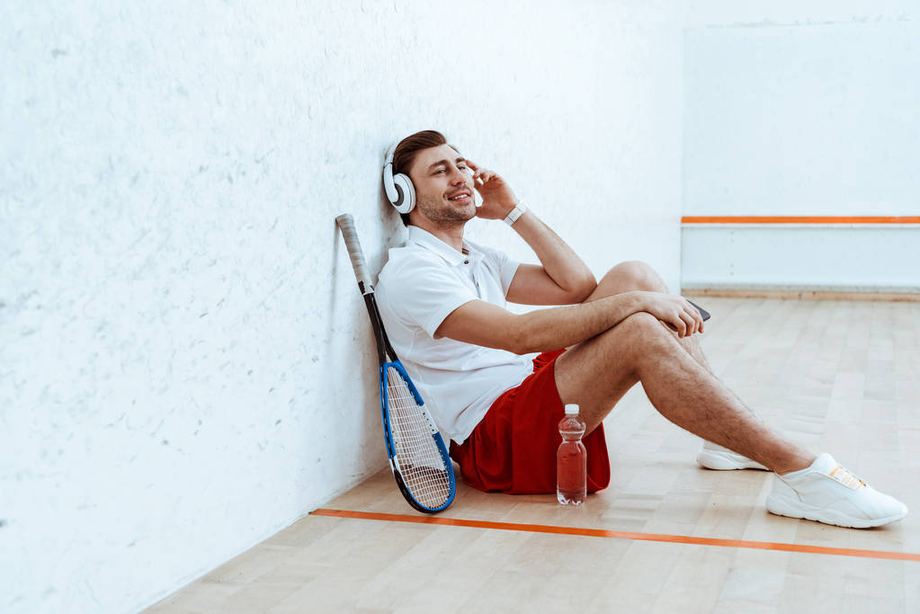 Χαμογελαστός παίκτης σκουός κάθεται στο πάτωμα και ακούγοντας μουσική στα ακουστικά - Φωτογραφία, εικόνα