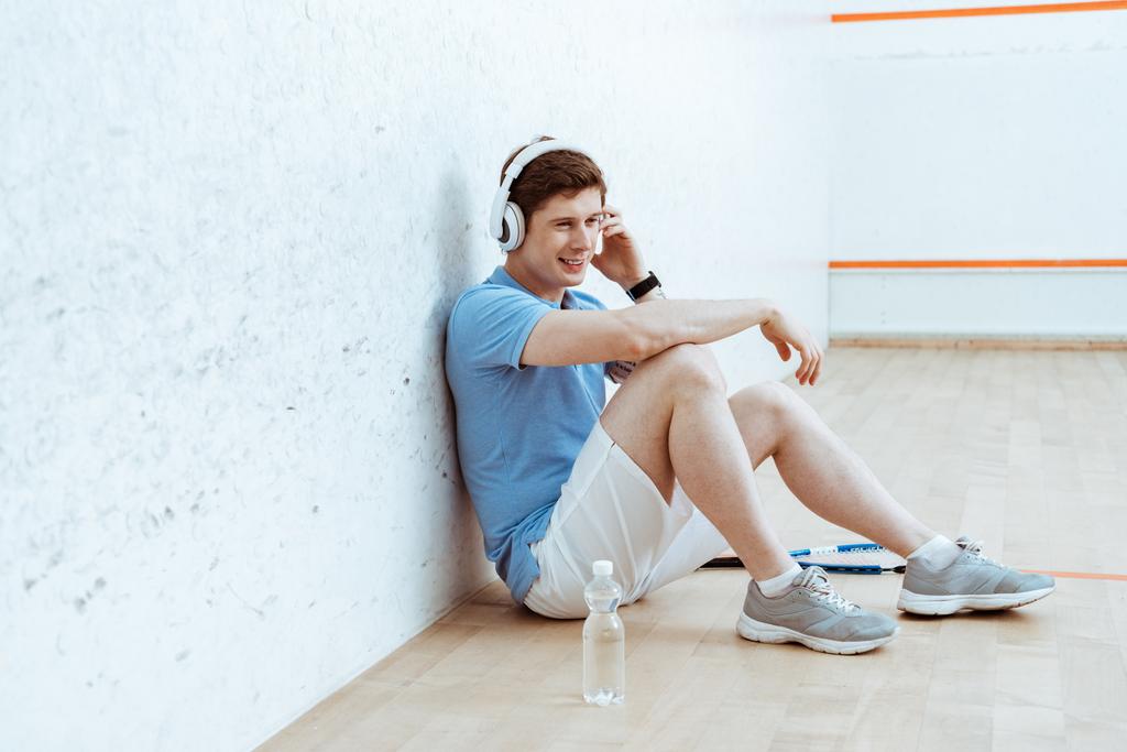 Lächelnder Sportler, der auf dem Boden sitzt und über Kopfhörer Musik hört - Foto, Bild