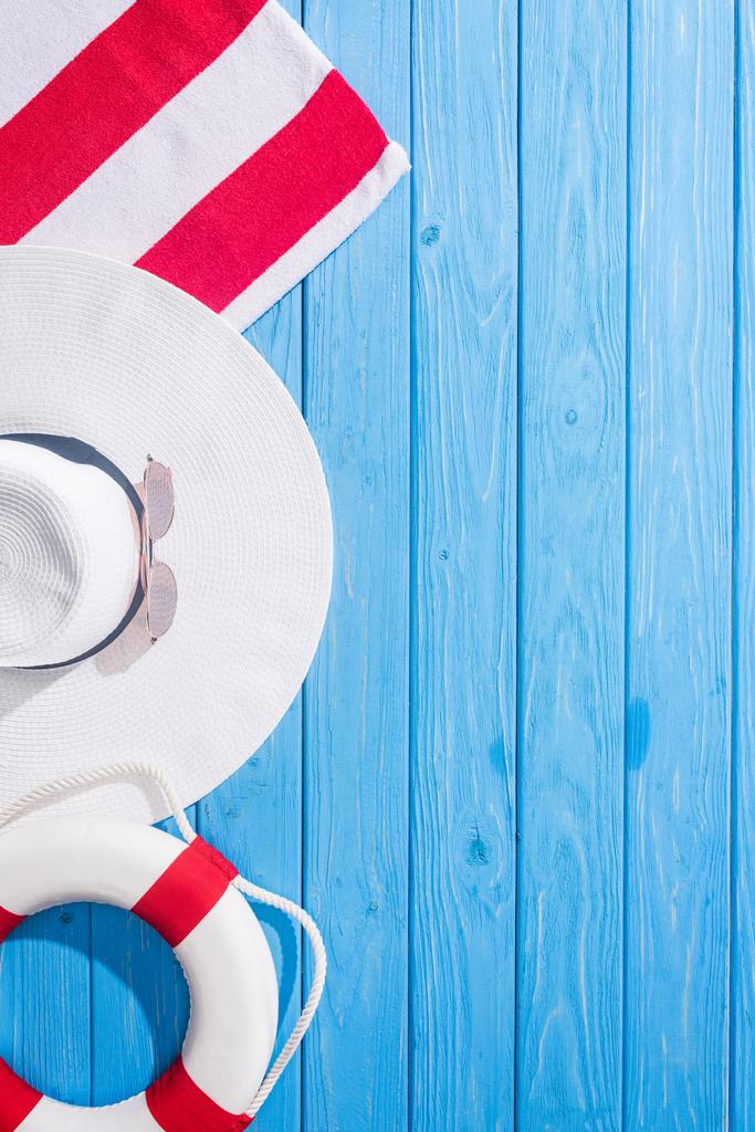 вид сверху на полосатое полотенце, белые солнцезащитные очки, спасательный круг, гибкая шляпа на синем деревянном фоне с копировальным пространством
 - Фото, изображение