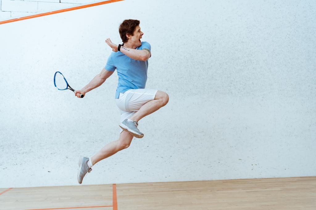 Urheilija poolopaidassa hyppii pelatessaan squashia neliseinäisessä tuomioistuimessa
 - Valokuva, kuva
