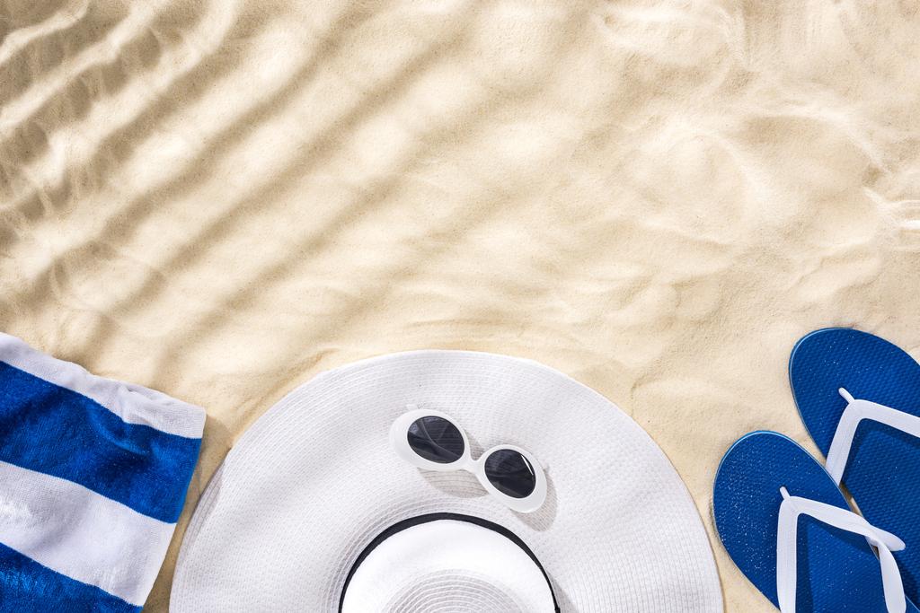 bovenaanzicht van gestreepte handdoek, retro zonnebril, floppy hoed en blauwe flip flops op zand met schaduw en kopieer ruimte - Foto, afbeelding