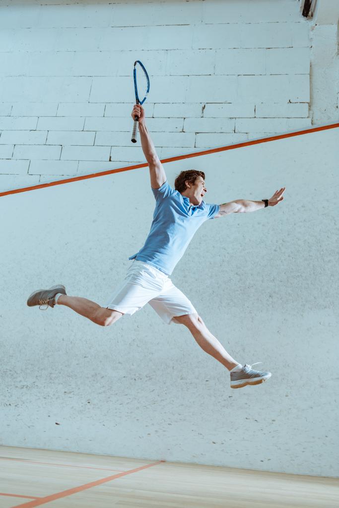 Vue complète du sportif excité en polo bleu sautant tout en jouant au squash
 - Photo, image