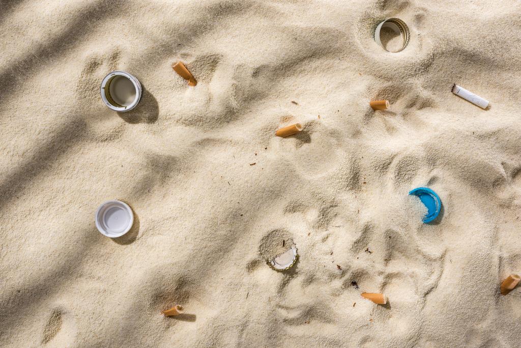 ylhäältä näkymä simpukat, pullonkorkit, hajallaan tupakantumppeja, muovipullo korkit hiekalla
 - Valokuva, kuva