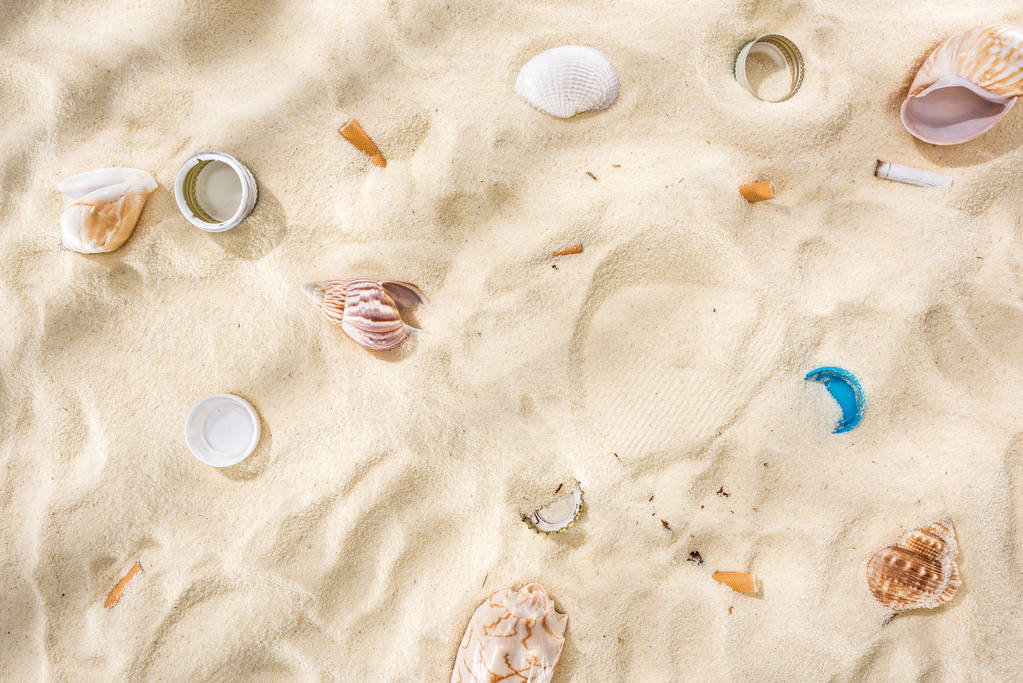 vista superior de conchas marinas, tapas de botellas, colillas de cigarrillos dispersas, tapas de botellas de plástico en la arena
 - Foto, Imagen