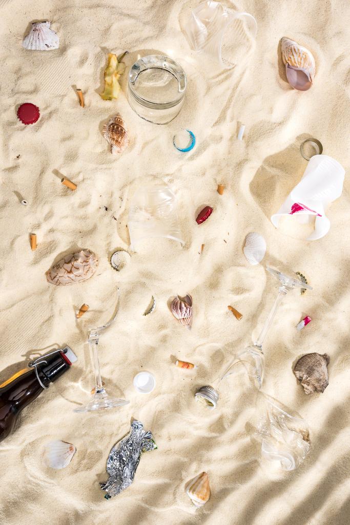 felülnézet a tengeri kagylókból, üvegpalackból, szétszórt cigarettacsikkek, törött szemüveg, Alma mag, műanyag csészék és cukorka wrapper homokon - Fotó, kép