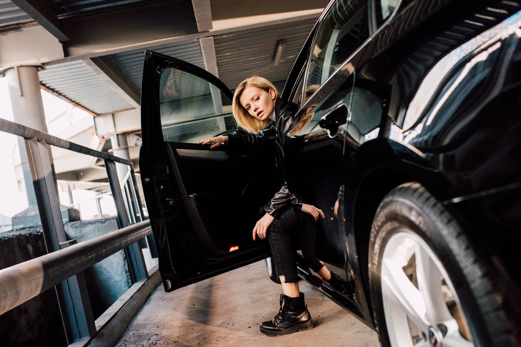 ελκυστική ξανθιά κορίτσι κάθεται στο αυτοκίνητο με ανοιχτή πόρτα και κοιτάζοντας την κάμερα  - Φωτογραφία, εικόνα