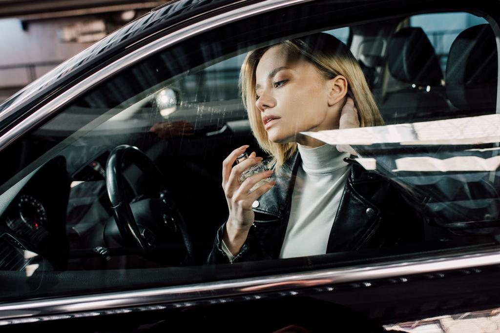 atrakcyjna blondynka trzyma butelkę podczas natryskiwania perfum w samochodzie  - Zdjęcie, obraz