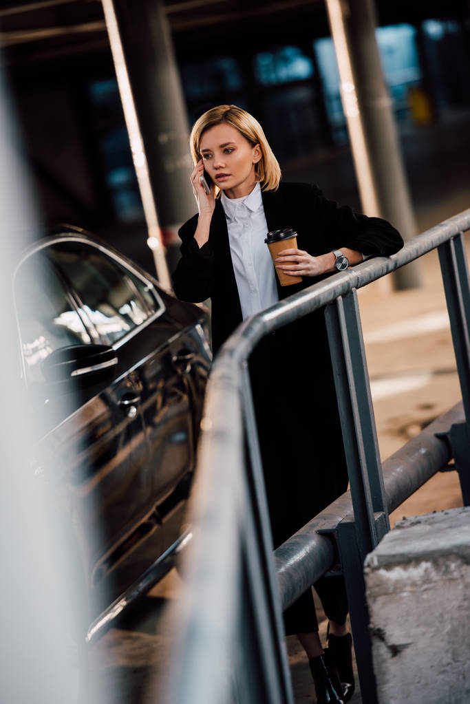 selective focus of worried blonde girl talking on smartphone and holding paper cup near black car - Photo, Image