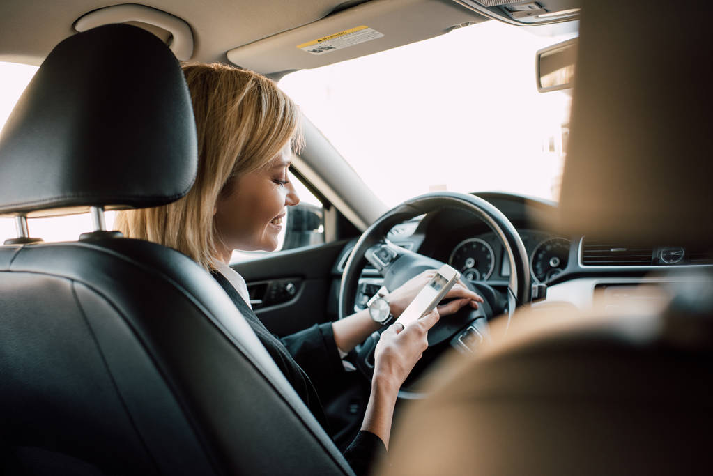 Wesoła blondynka młoda kobieta siedzi w samochodzie i za pomocą smartfona  - Zdjęcie, obraz