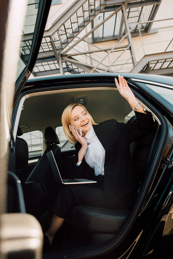 Tiefansicht einer glücklichen blonden Frau, die mit dem Smartphone spricht, während sie mit Laptop im Auto sitzt und mit der Hand winkt - Foto, Bild