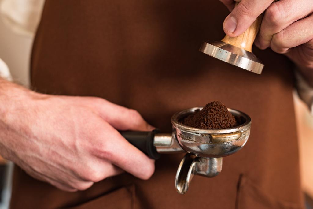 Bijgesneden weergave van Barista in bruin schort met filterhouder met gemalen koffie en knoeien - Foto, afbeelding