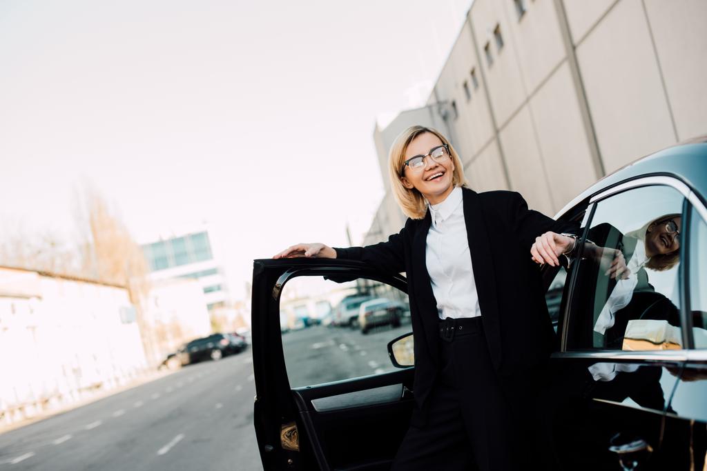 駐車場で黒い自動車の近くに立って眼鏡で幸せなブロンドの若い女性  - 写真・画像
