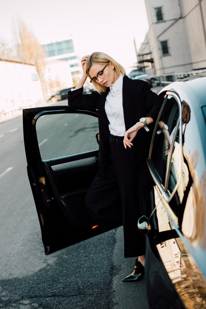 Blondýnka v brýlích stojících v blízkosti černého automobilu a dotýkání se vlasů   - Fotografie, Obrázek