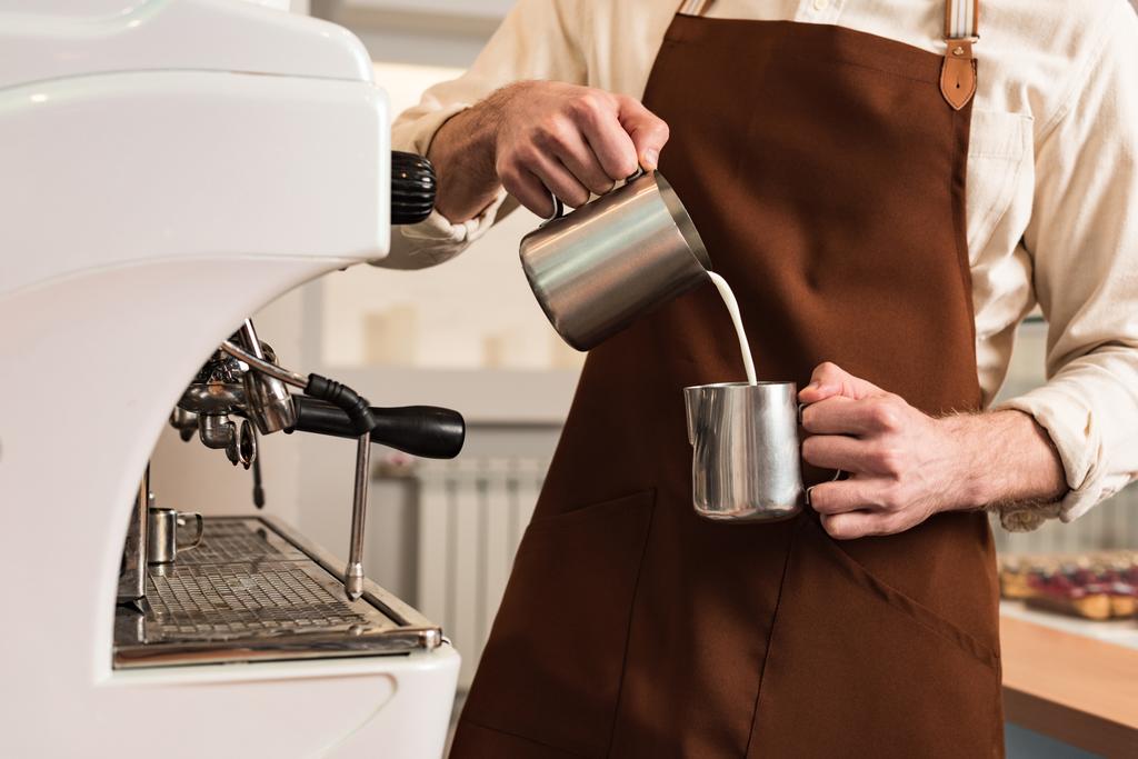 Vista ritagliata di barista in grembiule marrone versando latte in tazza d'acciaio
 - Foto, immagini